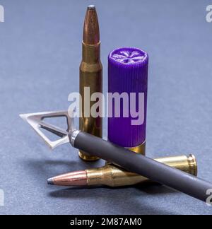 An arrow with a three blade broadhead attached to it with two 30.06 caliber bullets and a 12 gauge shotgun shell on a gray background Stock Photo