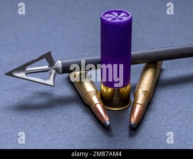 An arrow with a three blade broadhead attached to it with two 30.06 caliber bullets and a 12 gauge shotgun shell on a gray background Stock Photo