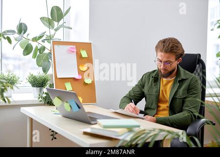 Nice man office worker concentrated on work, writing documents information, sit alone in bright cozy modern office, use laptop, dressed in casual clot Stock Photo