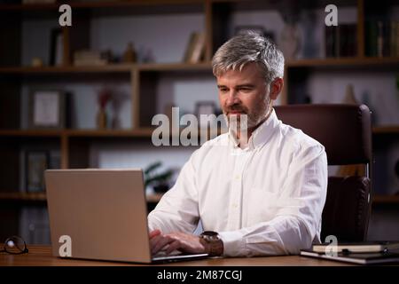 Middle-aged man freelancer in eyeglasses working on laptop remotely from office. Serious businessman chatting online typing on a laptop keyboard Stock Photo