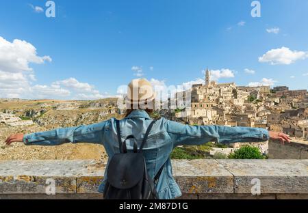 Young girl with hat standing with open arms looking at the ancient city of Matera on a sunny day, Italy. Stock Photo