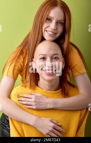 Two excited ladies with red and bald hair head looking at camera posing, hugging together, isolated over green studio background. Portrait of cheerful Stock Photo