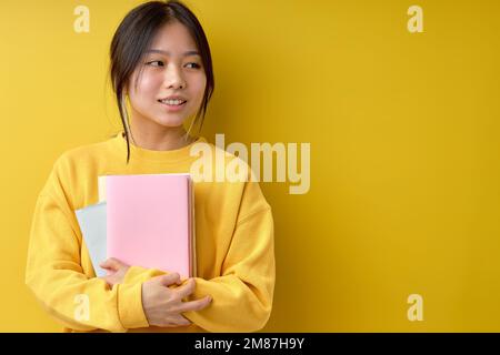 Pretty cheerful attractive beautiful young brunette asian woman 20s wearing basic casual yellow shirt standing with books, looking at side, isolated o Stock Photo