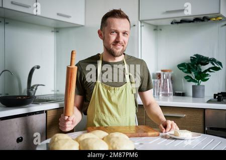 Caucasian baker man portrait in green apron uniform smiling and looking at camera holding rolling pin. Handsome male baker at home sitting at the table posing. High quality photo Stock Photo