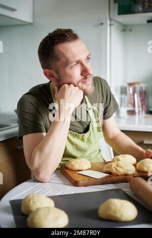 Attractive caucasian male chef baker in green apron thinking about homemade pastry. Dough balls proofing on cutting board. Working at home kitchen concept, homemade baking. High quality vertical image Stock Photo