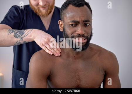 cropped caucasian Spa therapist making relaxing massage for handsome middle aged black man, rubbing back. Frustrated bearded man getting healing spine Stock Photo