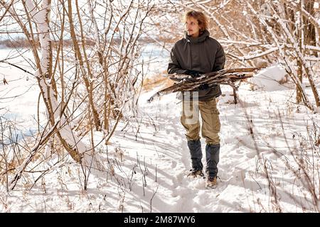 young caucasian male gathering wood for fire in forest, nature. lonely guy in warm clothes in snowy winter forest. bushcraft, camping, travel, active Stock Photo