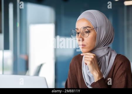 Close up of thinking business woman in hijab in modern office, serious muslim woman looking at laptop screen thinking about decision, woman at workplace successful. Stock Photo