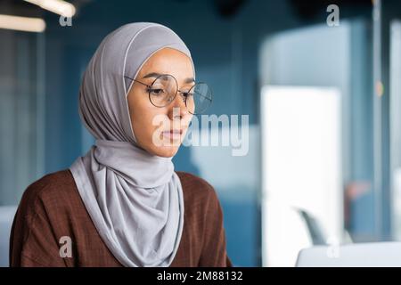 Close up of thinking business woman in hijab in modern office, serious muslim woman looking at laptop screen thinking about decision, woman at workplace successful. Stock Photo