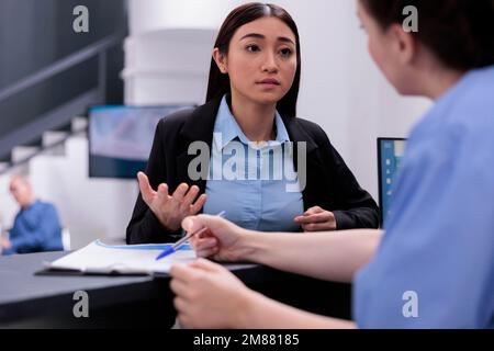 Assistant showing medical report to receptionist discussing expertise during checkup visit examination in hospital waiting area. Worker helping nurse with consultations and follow-up appointments. Stock Photo