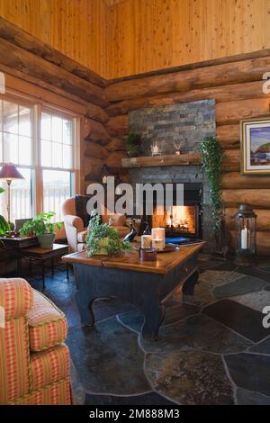 Upholstered armchairs and wooden coffee table with lit natural stone fireplace in living room inside handcrafted Scandinavian spruce log home. Stock Photo