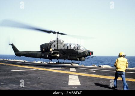 An AH-1 Sea Cobra helicopter takes off from the flight deck of the amphibious assault ship USS GUALDALCANAL (LPH-7) as a landing signal officer stands by. Country: Gulf Of Oman Stock Photo