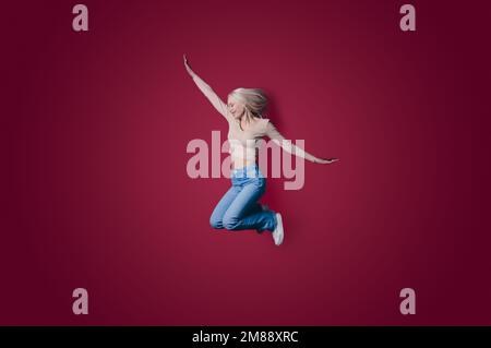 Full size photo of cool blonde woman jumping wearing jeans isolated on cherry-colored background Stock Photo