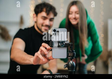 Smiling young couple with happy faces looking at phone camera, bloggers recording videoblog at home. Video live streaming. Stock Photo