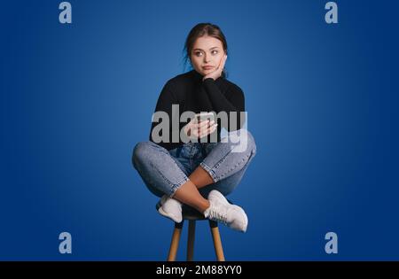 Portrait of attractive minded girl sitting on chair using phone chatting isolated overblue color background Stock Photo