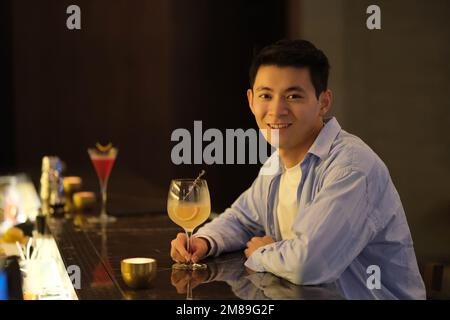 Asian young man sit at night bar counter with a glass of cocktail, smile at camera Stock Photo