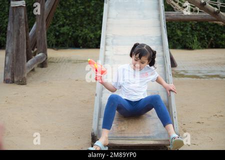 Happy child girl has fun playing sliding and shooting water gun on outdoor playground on hot summer day outdoors. Stock Photo