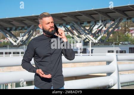 Middle-aged man with a dark beard and blond hair combed back of his head, in a black shirt, is talking on the phone on a city street near the railway Stock Photo