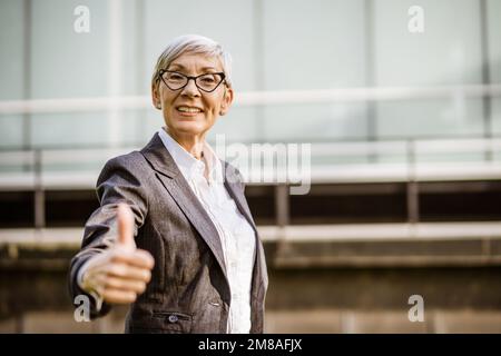 Outdoor portrait of successful mature businesswoman in front of company building. She is looking at camera and showing thumbs up. Stock Photo