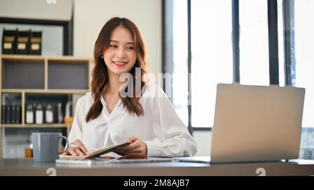 Beautiful and charming millennial Asian businesswoman or female secretary working at her desk in the office, looking at laptop screen, checking her bo Stock Photo