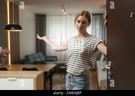 Cheerful woman opening house front door and inviting friends. Young woman welcoming visitors in her home. Stock Photo