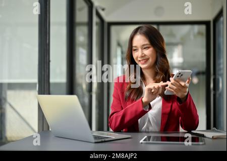 Attractive millennial Asian businesswoman using her modern smartphone while looking at laptop screen at her desk. Stock Photo
