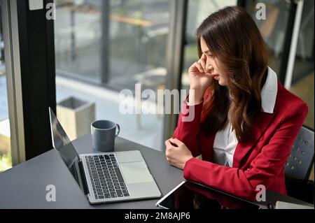 Stressed and thoughtful millennial Asian businesswoman looking at laptop screen, working at her desk. Stock Photo