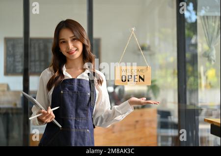 Beautiful and friendly young Asian female waitress or coffee shop staff, in an apron with her tablet, stands at the entrance door of her cafe, welcomi Stock Photo