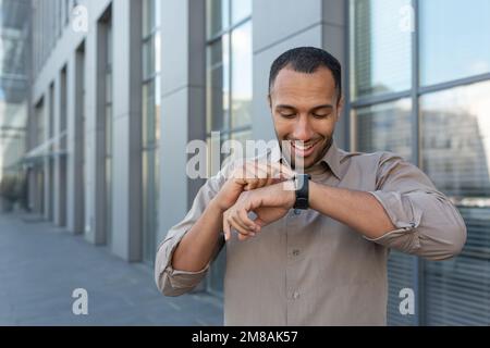 Latin American businessman in shirt cheerful talking with friends using smartwatch, man outside office building using gadget for artificial intelligence commands. Stock Photo