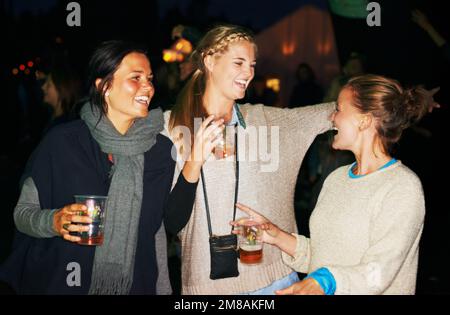 Young and not a care in the world. three friends talking and drinking at night at an outdoor music festival. Stock Photo