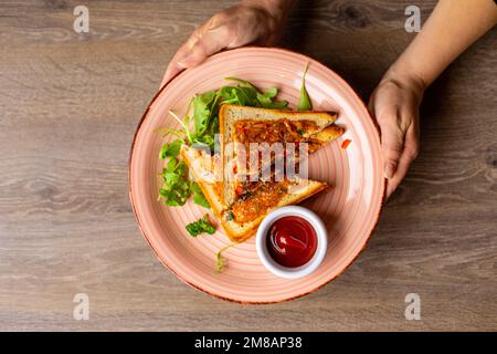 Cropped photo of woman holding pink plate with sandwiches, ketchup in bowl decorated with greenery on wooden table. Stock Photo