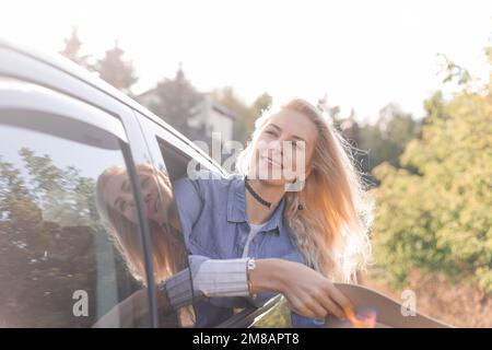 Glad, pensive, smiling, proud blond woman hold hat in hands, leaning out of car window on backseat. Journey, travelling Stock Photo
