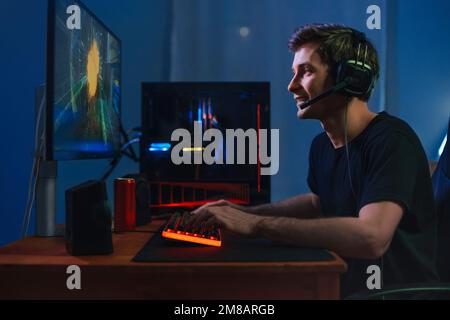 Young handsome pro gamer playing in online video game on computer at home using headphones and microphone for talking with his team, smiling, enjoying the play. Side view. Cybersport concept Stock Photo