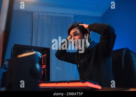 Portrait of young male cyber sport player shocked with winning online tournament or shooter video game, keeps hands on his head, feel exited. Side view, neon light, close up. Professional cyber sport Stock Photo