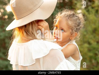 Portrait closeup of cute curly little girl sitting on hands of mother standing with back in hat. Mom and child in forest at summer evening, family Stock Photo