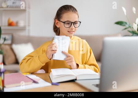 School girl wearing eyeglasses having video call with online tutor, using laptop for distance learning with teacher, showing him exercise via webcam, studying from home. Homeschooling due to covid-19 Stock Photo