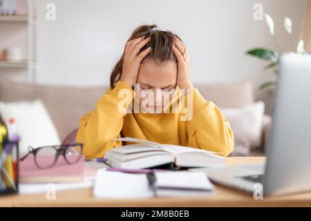Exhausted schooler girl suffering from doing homework, tired from studying at home, getting ready before exams, stressed with difficult task, reading book, using laptop. Home education, homeschooling Stock Photo