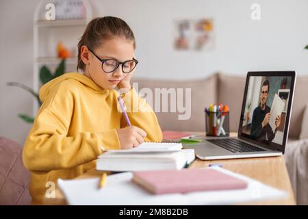 Tired girl in eyeglasses having online lesson with personal tutor on laptop. Kid bored from studying, writing math exercise in notebook. Side view. Elearning and remote school during covid quarantine Stock Photo
