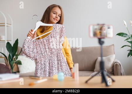 Young blogger girl talking about fashion on smartphone camera, presenting dress, shooting vlog for her followers at home.Popular kid influencer recording video podcast about stylish clothes. Blogging Stock Photo