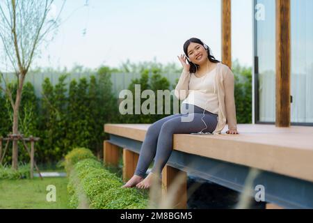 Portrait of pregnant Happy Asian woman sitting on terrace at home and using smart phone for listening music in headphones. Stock Photo