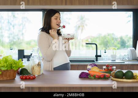 Beautiful Asian pregnant woman eating with lust cereals on breakfast in kitchen at home, enjoying meal. Yummy food for pregnant Stock Photo