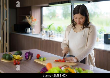 Pregnant Asian woman cutting tomato for fresh green salad, female prepares tasty organic dinner at home, healthy nutrition for future mother Stock Photo