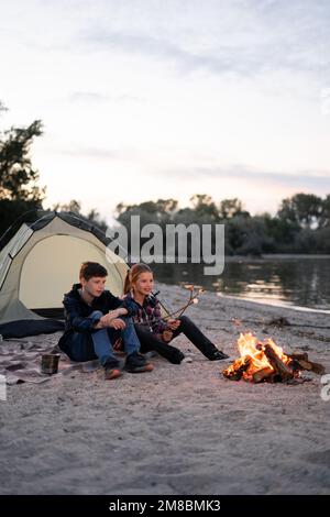 happy kids roasting bbq marshmallows on sticks around campfire resting at campsite Stock Photo