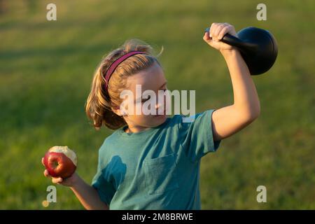 Sporty kid with apple and kettlebell outdoor in summer park. Sport activities at leisure with children. Blonde boy holding kettlebell. Sports Stock Photo