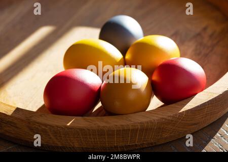 six brightly colored Easter eggs lie on a wooden tray in the sun Stock Photo