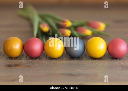 colored Easter eggs lie in a row on a wooden table, next to tulips Stock Photo
