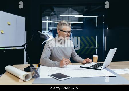 Senior gray-haired man architect, engineer, designer sits in the office at the table, works on a laptop, drawings. Makes notes. Stock Photo