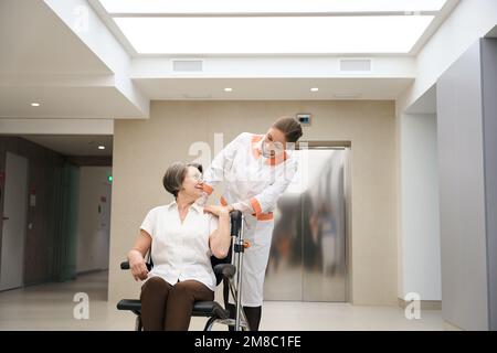Medical worker is carrying patient in a wheelchair through foyer Stock Photo