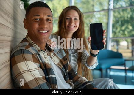 Pretty woman holding phone and sitting next to her male colleague Stock Photo