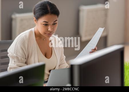 focused multiracial businesswoman holding document while working on laptop near blurred computer monitors,stock image Stock Photo